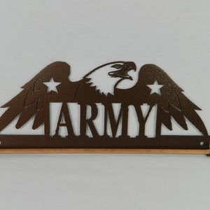 May include: A brown metal wall decor with a silhouette of an eagle with wings spread and the word "ARMY" in the center. The eagle has two stars on its wings. The decor is attached to a wooden dowel rod with wooden finials.