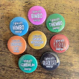 Op de afbeelding: Zeven ronde knoppen in verschillende kleuren met tekst. Elke knop zegt "I give off" gevolgd door een ander woord zoals "Bimbo", "Himbo", "Thembo", "Orange Cat", "Golden Retriever", "Feral", "Gremlin" en "Black Cat". Alle knoppen hebben het woord "energy" onderaan.