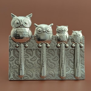 May include: Set of four silver owl-shaped measuring spoons with a paisley patterned holder. The spoons are labeled "tablespoon", "teaspoon", "half teaspoon", and "quarter tsp".