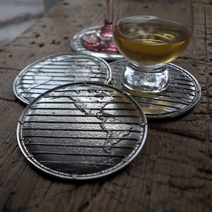May include: Four silver coasters with a world map design. The coasters are round and have a raised edge. The coasters are arranged in a circle with a glass of amber liquid in the center.