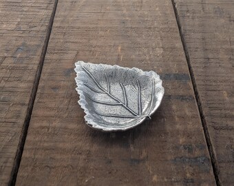 May include: A silver leaf-shaped dish with a textured surface. The dish is made of metal and has a natural leaf design.