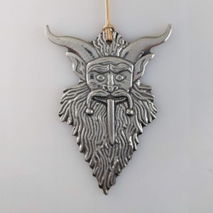May include: A silver-colored metal ornament featuring a horned, bearded face with a long tongue. The ornament has a textured surface and is suspended by a thin, tan string. The design is detailed and stylized, with a focus on the face's features.