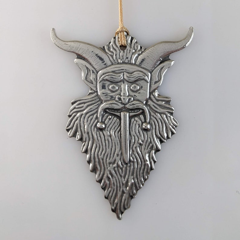 May include: A silver-colored metal ornament featuring a horned, bearded face with a long tongue. The ornament has a textured surface and is suspended by a thin, tan string. The design is detailed and stylized, with a focus on the face's features.