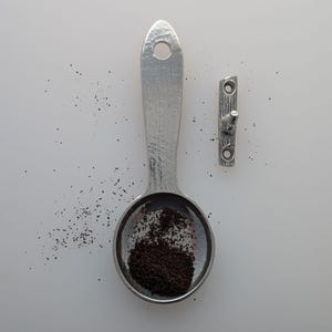 Könnte beinhalten: Ein silberfarbener Metalllöffel gefüllt mit dunkelbraunem Kaffeepulver, neben einem kleinen, passenden Metallhaken. Der Löffel hat einen langen Griff mit einem Loch oben. Der Haken hat ein strukturiertes, astähnliches Design.