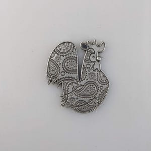 Peut inclure: Une figurine de coq en argent avec un motif paisley. Le coq est debout sur une surface blanche.