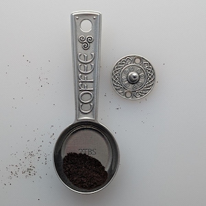 Könnte beinhalten: Ein silberner Kaffeelöffel mit dem Wort "COFFEE" auf dem Griff eingraviert. Der Löffel enthält gemahlenen Kaffee und ist mit "2TBS" beschriftet. Ein dekorativer silberner Knopf mit einem keltischen Knotenmuster ist ebenfalls vorhanden.