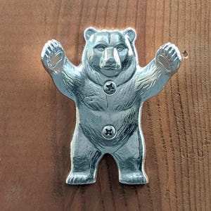 May include: A silver-toned metal wall hook shaped like a standing bear with its arms raised. The bear figure features detailed fur texture and is secured to the surface with two visible Phillips head screws positioned on its chest and abdomen.