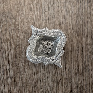 Puede incluir: Un pequeño y ornamentado plato de plata con un detallado diseño floral. El plato tiene forma de cuadrifolio con un área cuadrada central. La superficie tiene una textura martillada y los bordes están ligeramente elevados.