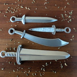 May include: Four silver-colored miniature swords with detailed handles and blades are arranged on a wooden surface. The swords vary in shape, including a straight blade, a curved blade, and a dagger-like design. Dried herbs are scattered around the swords.