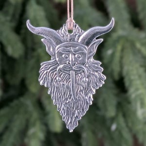 May include: A silver-colored ornament featuring a horned face with a long beard and a stern expression. The ornament is suspended by a tan string, and the background is a blurred view of evergreen branches. The ornament is likely for holiday decor.
