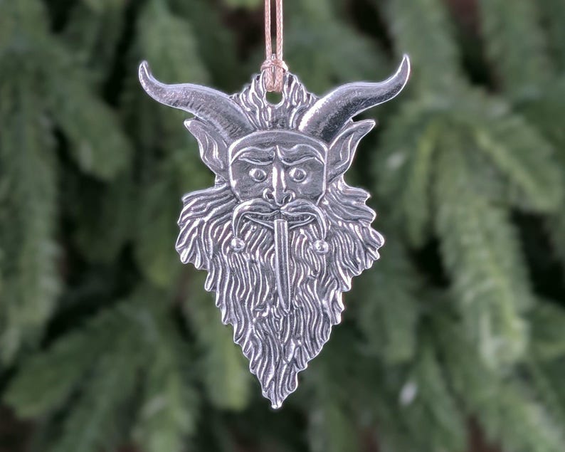 May include: A silver-colored ornament featuring a horned face with a long beard and a stern expression. The ornament is suspended by a tan string, and the background is a blurred view of evergreen branches. The ornament is likely for holiday decor.