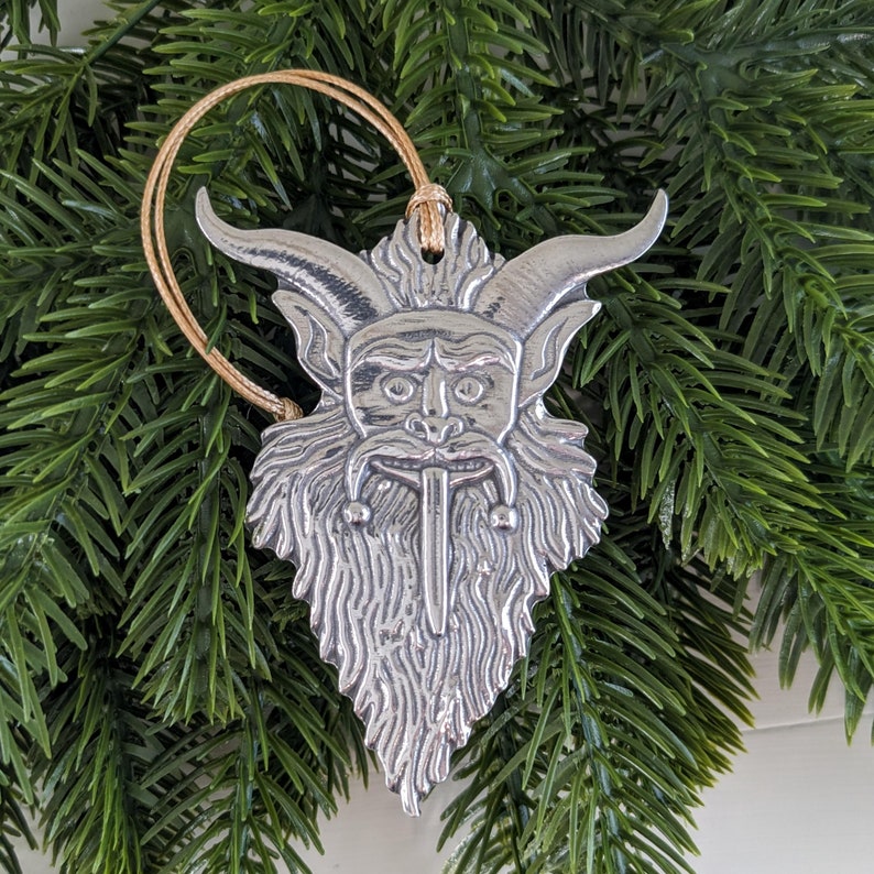 May include: A silver-colored Christmas ornament featuring a Krampus design. The ornament has a detailed face with horns, a long tongue, and a beard. It hangs from a beige string against a backdrop of green pine needles.