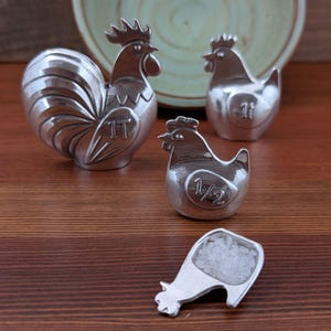 May include: A set of silver-colored rooster and hen salt and pepper shakers. The rooster and hens have the numbers "1T" and "1/2" on them. The salt shaker is shaped like a hen and is filled with white salt.