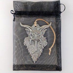 May include: A silver-colored pendant in the shape of a horned, bearded face with a long, pointed beard, hanging from a tan string. The pendant is inside a black mesh drawstring bag.