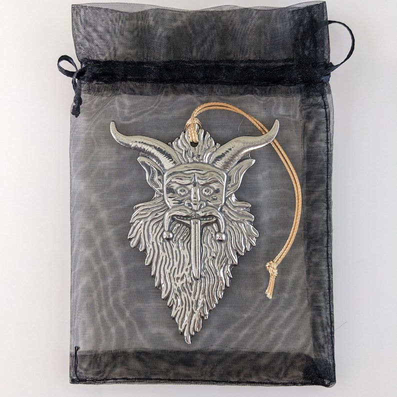 May include: A silver-colored pendant in the shape of a horned, bearded face with a long, pointed beard, hanging from a tan string. The pendant is inside a black mesh drawstring bag.