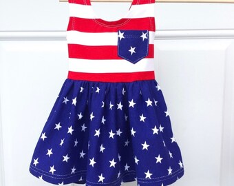 May include: A sleeveless dress featuring a bodice with horizontal red and white stripes and a single chest pocket. The skirt is blue with a white star pattern. The pocket is also blue with white stars. The dress is displayed on a wooden hanger.