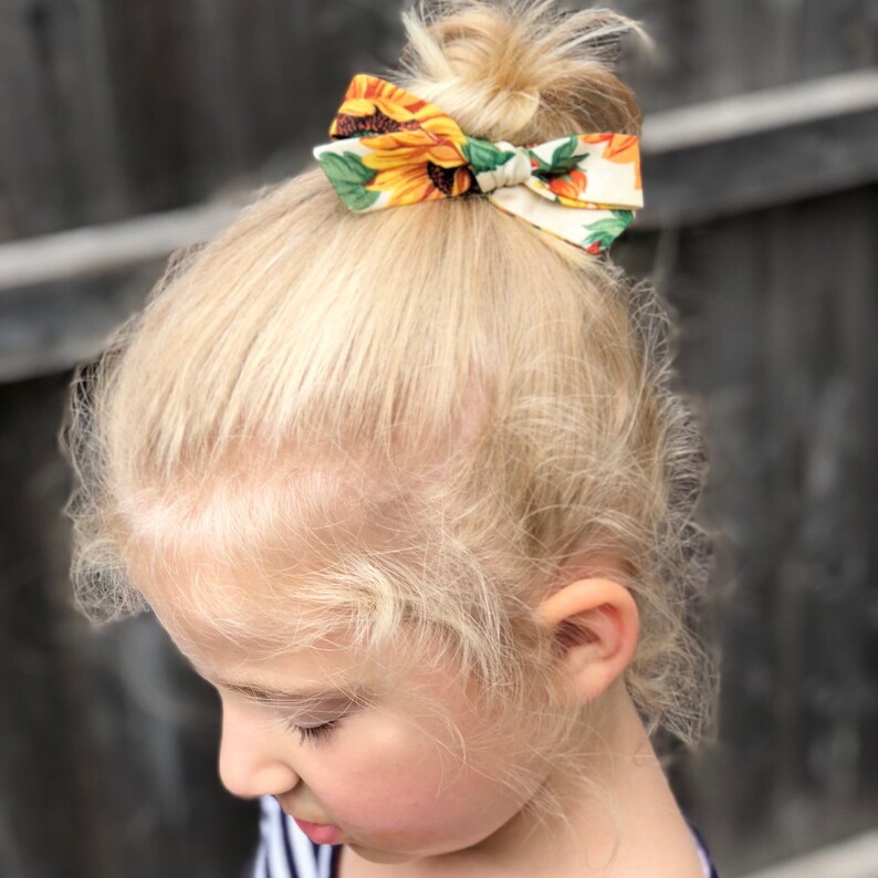 Op de afbeelding: Een jong meisje met blond haar draagt een haarband met een zonnebloemprint. De band is bovenop haar hoofd in een knoop geknoopt.