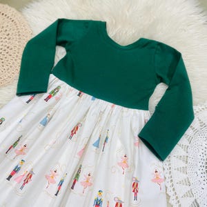 May include: A long-sleeved dress with a solid dark green bodice and a white skirt featuring a repeating pattern of nutcracker figures, ballerinas, and mice in pink, blue, and red, accented with thin gold swirling lines throughout the fabric.