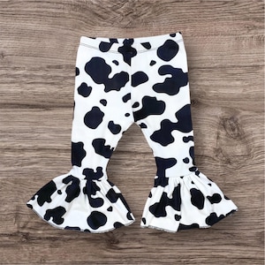 May include: Black and white cow print bell bottom pants for a baby or toddler. The pants have a stretchy waistband and a ruffled hem.