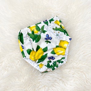May include: White baby bloomers with a lemon and blueberry print. The bloomers have a white elastic waistband and are made of a soft, comfortable fabric.