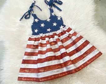 May include: A red, white, and blue sleeveless dress with a ruffled skirt. The top of the dress features a blue background with white stars, while the skirt has red and white stripes with a woodgrain pattern.