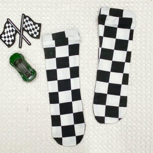 May include: Two black and white checkered socks are displayed on a white surface, along with a green toy car and two checkered racing flags. The socks have a classic racing flag pattern.