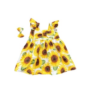 May include: A white dress with a yellow sunflower print, featuring a matching bow headband. The dress has a square neckline, ruffled straps, and a bow detail at the chest. The sunflowers are in full bloom, with yellow petals and brown centers.