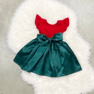 Op de afbeelding: Een rood en groene jurk met een grote groene strik. De jurk heeft korte mouwen en een volle rok.