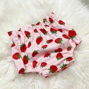 May include: Pink baby bloomers with a strawberry print. The bloomers have a white polka dot pattern and are made of soft cotton.