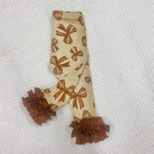 May include: Cream-colored leggings with a repeating pattern of brown football-shaped bows. The leggings have ruffled brown trim at the ankles. The fabric has a ribbed texture.