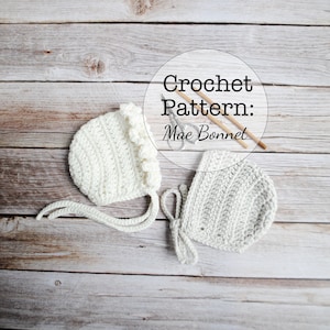 May include: Two crocheted baby bonnets, one white and one gray, are displayed on a wooden surface. The white bonnet has a ruffled edge. The image includes the text "Crochet Pattern: Mae Bonnet" in a circular design, along with knitting needles and scissors.