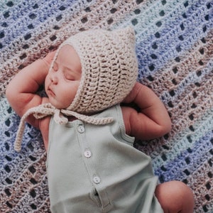 May include: A sleeping baby wearing a light beige crochet bonnet with ear flaps and a light green ribbed romper. The bonnet has tie closures. The baby is lying on a multi-colored, open-weave blanket with shades of blue, brown, and beige.
