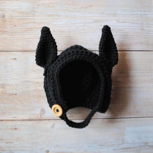May include: A black crochet hat with tall, pointed ears and a wooden button closure. The hat is designed to cover the head and has a loop for securing under the chin. The hat is made of a textured yarn.