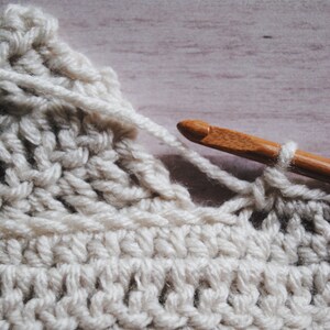 Pu&ograve; includere: Primo piano di un tessuto all'uncinetto con un uncinetto di legno. Il tessuto &egrave; di colore crema chiaro e l'uncinetto viene utilizzato per creare un punto.