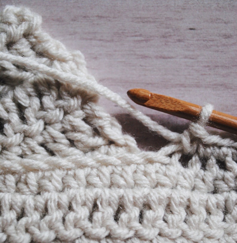 Pu&ograve; includere: Primo piano di un tessuto all'uncinetto con un uncinetto di legno. Il tessuto &egrave; di colore crema chiaro e l'uncinetto viene utilizzato per creare un punto.