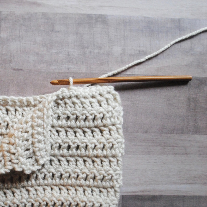 Pu&ograve; includere: Primo piano di un pezzo all'uncinetto di colore crema, con un uncinetto di legno e filo. L'uncinetto viene utilizzato per lavorare un punto nel tessuto.