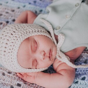 Pu&ograve; includere: Un bambino che dorme con un berretto di uncinetto bianco con un lungo laccio, sdraiato su una coperta di uncinetto blu e bianca.