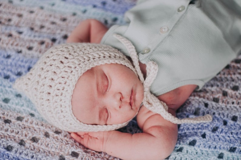 Pu&ograve; includere: Un bambino che dorme con un berretto di uncinetto bianco con un lungo laccio, sdraiato su una coperta di uncinetto blu e bianca.