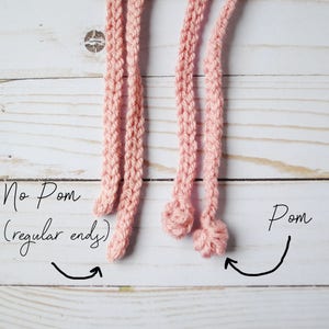 Pu&ograve; includere: Due cordoni di uncinetto rosa, uno con un'estremit&agrave; normale e l'altro con un'estremit&agrave; a pompon. Il testo "No Pom (regular end)" &egrave; scritto accanto al cordone con un'estremit&agrave; normale, e il testo "Pom" &egrave; scritto accanto al cordone con un'estremit&agrave; a pompon.