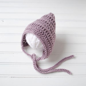Pu&ograve; includere: Un berretto per neonato in crochet color malva con un lungo laccio. Il berretto &egrave; realizzato con un filato morbido e testurizzato e ha una cima a punta.