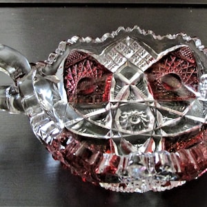 Könnte beinhalten: Eine klare Glasschale mit roter Tönung und einem geschliffenen Glasdesign. Die Schale hat einen einzigen Griff.