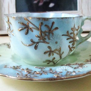 May include: A vintage teacup and saucer set with a light blue and green glaze. The set is decorated with gold leaf floral designs.