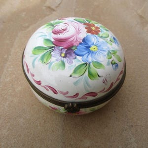 May include: A white porcelain trinket box with a floral design. The lid is decorated with hand-painted pink, blue, purple, and green flowers. The box has a brass hinge and a brass clasp.