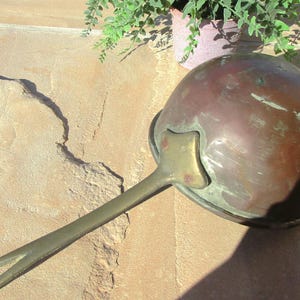 May include: A vintage copper ladle with a long, brass handle. The ladle has a rounded bowl and a decorative brass accent on the handle.