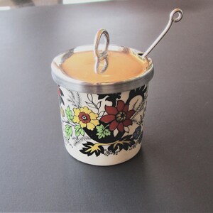 May include: A white ceramic jar with a floral design and a silver lid. The jar has a silver spoon attached to the lid.