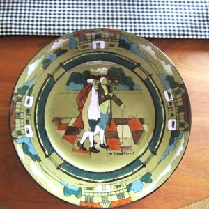May include: A vintage ceramic plate with a green, white, and brown design. The plate features a scene of two men walking on a checkered path, with buildings in the background. The text "Ye Village Street" is written on the plate.