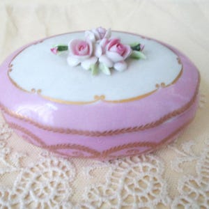 May include: A small, oval-shaped porcelain trinket box. The box is light pink with a white top and gold trim. The lid is adorned with a cluster of pink and white porcelain roses and green leaves. The box is likely vintage.