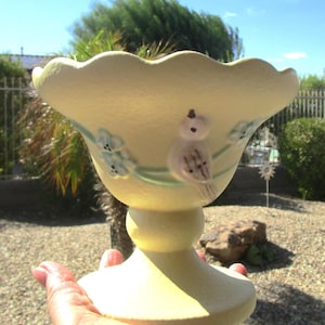 May include: A yellow ceramic pedestal bowl with a scalloped rim and a bird design. The bird is painted in shades of pink, blue, and brown. The bowl is decorated with blue flowers.