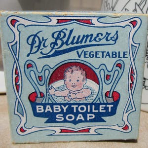 May include: A blue and white box with a red and white border. The box has a picture of a baby in a bathtub and the text "Dr. Blumors Vegetable Baby Toilet Soap".