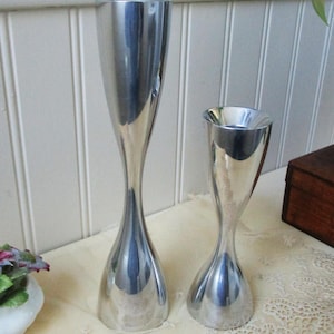 May include: Two polished silver candle holders with a sleek, modern design. The taller holder is approximately 12 inches tall, while the shorter one is about 7 inches. Both have a flared top and a bulbous base, reflecting the surrounding light.